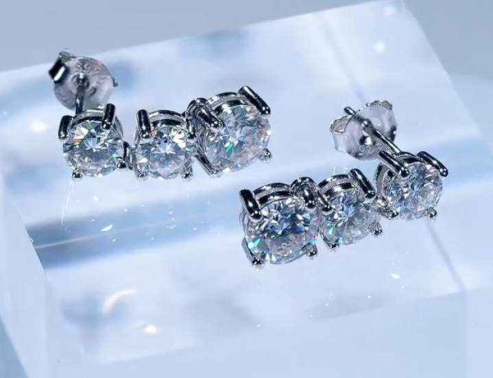 Pair of round-cut moissanite earrings set in rhodium-plated sterling silver, displayed on a clear ice-like surface with crisp brilliance and clean reflections.