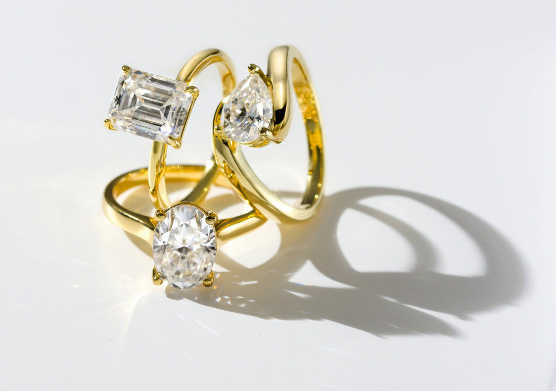 Goldene Ringe mit Moissanit-Steinen in verschiedenen Schliffen (Oval, Emerald, Herz) auf hellem Hintergrund mit weichen Lichtreflexionen.